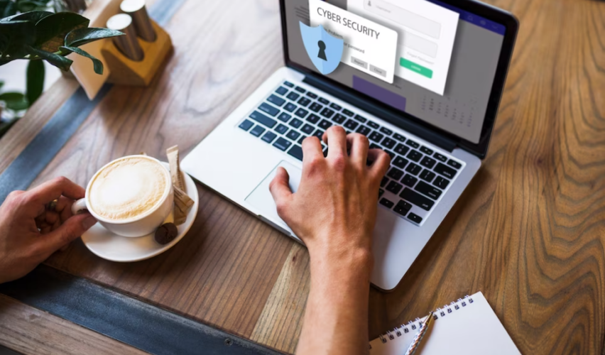 hand typing on a laptop, other holds a coffee, cyber security on the laptop screen